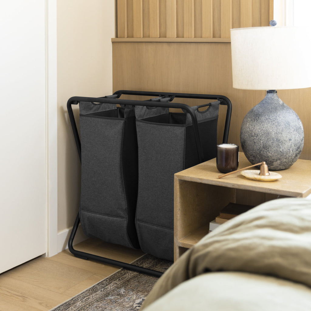 Angled lifestyle view of Simplehuman Double X-Frame Hamper, Matte Black, dual removable sorting liners in a bedroom.