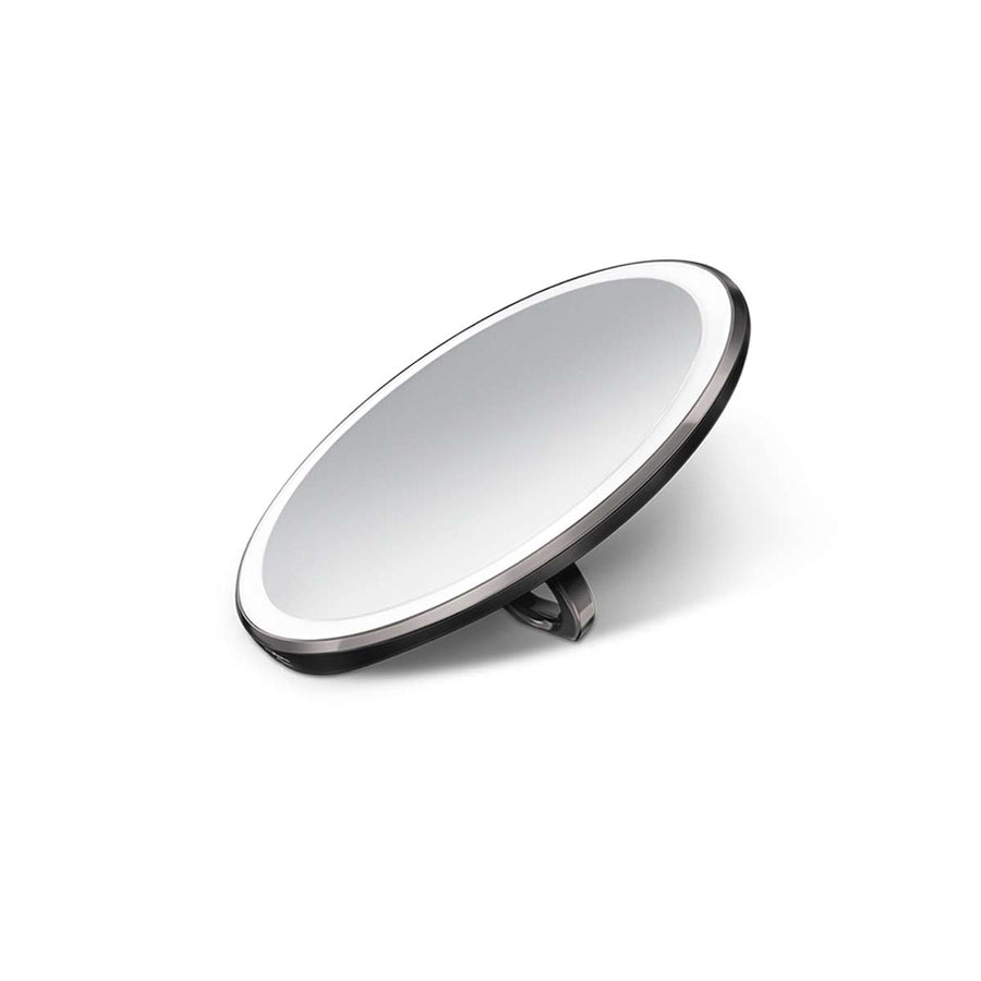 Angled view of Simplehuman Sensor Mirror Compact 3x in Matte Black, illuminated, propped open for hands-free use.
