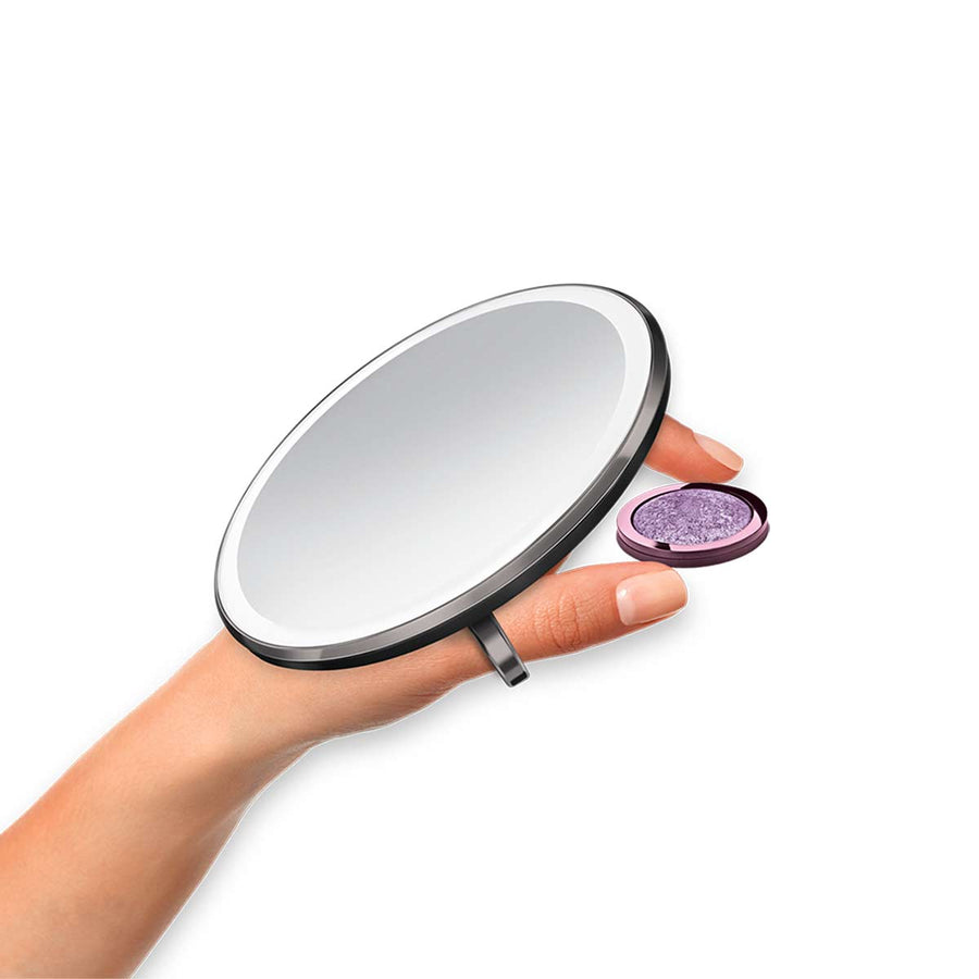 Angled view of Simplehuman Sensor Mirror Compact 3x in Matte Black, held using the finger ring for secure makeup application.