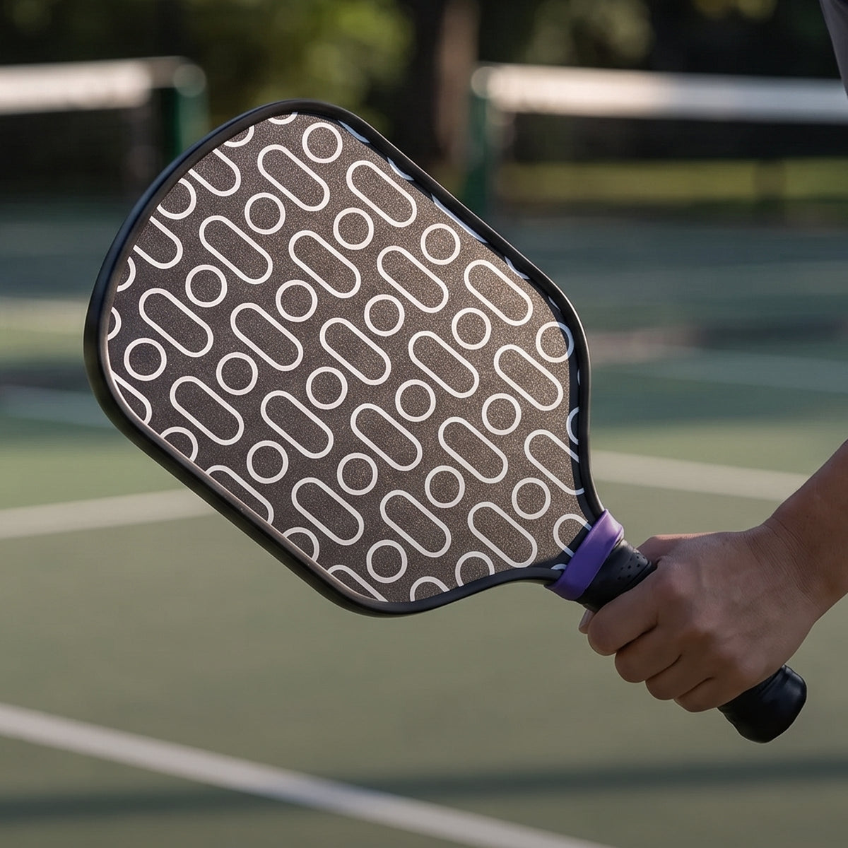 A close-up of a pickleball paddle being held by a person on an outdoor pickleball court. The paddle has a dark gray textured face with a repeating white abstract pattern, a black frame, and a purple grip accent.
